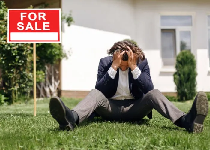 Depressed real estate agent sitting on grass in front of house for sale, outside as an image for a piece on For Sale by Owner Homes