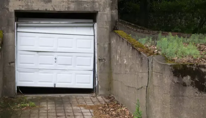 A single garage door hangs askew and dented in a property that appears, dirty, neglected and covered in old leaves