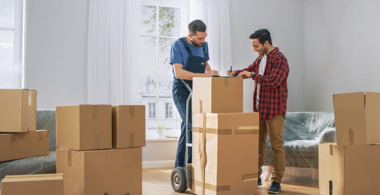 Happy New Homeowner Welcomes Professional Mover with Hand Truck full of Cardboard Boxes, Receives His Goods and Signs on Clipboard as an image for a piece on the best moving companies