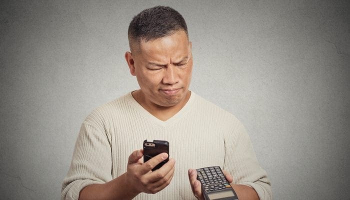 Confused man looking at his smart phone holding calculator isolated on gray wall background as an image for a piece on what is a pocket listing