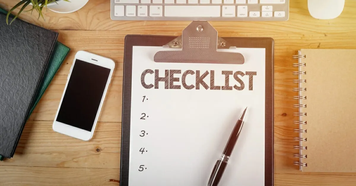 A phone and a clipboard sit on a modern wooden desk, and a paper with the words CHECKLIST sits on the clipboard next to a black pen