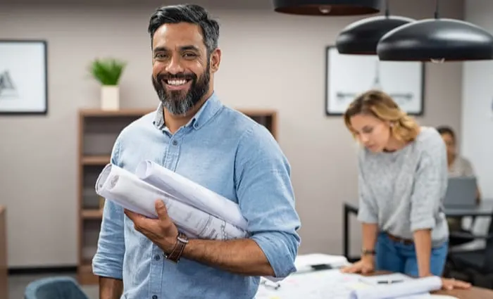 Portrait of multiethnic architect with blueprints in creative office. Mature middle eastern contractor holding roll of architectural projects while looking at camera. Happy latin man in casual.