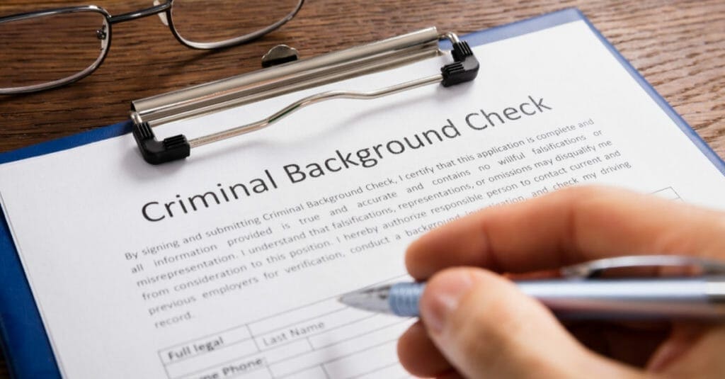A person holding a blue pen filling out a criminal background check form with glasses sitting on the table