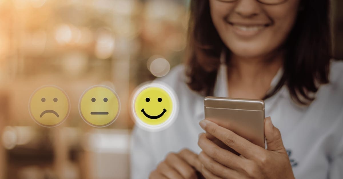 A business woman holding a mobile phone and tapping the smiley face emoji to symbolize a positive customer review