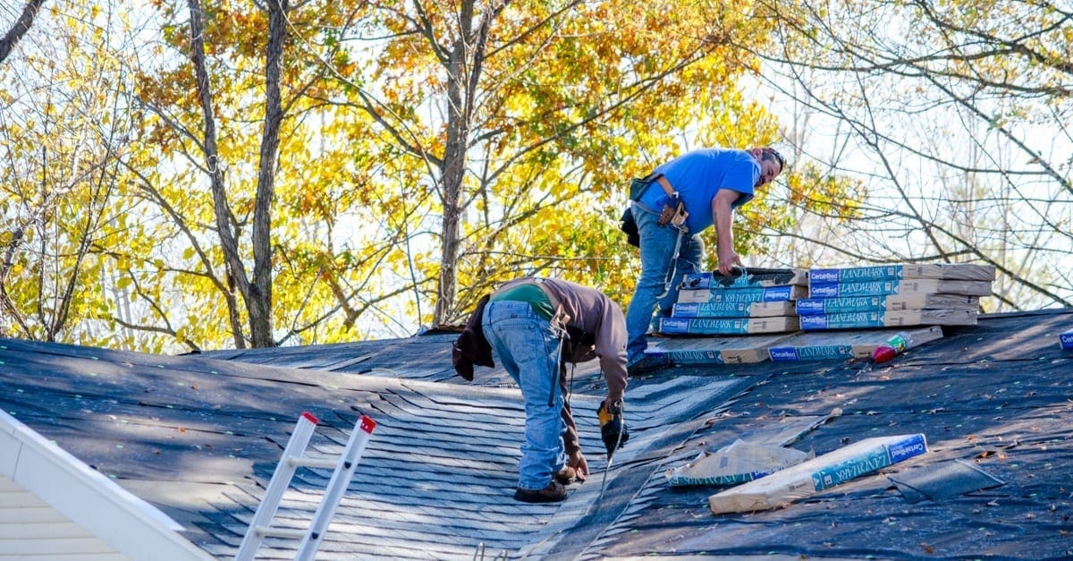 Two workers on a roof with materials and trees in the background as the featured image for a piece on how to get insurance to pay for roof replacement