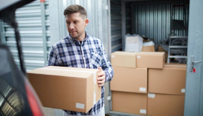 As an image for a piece on moving container companies, Serious handsome young man in casual clothing finding place in car for cardboard box while loading automobile at container storage area, he storing things in container