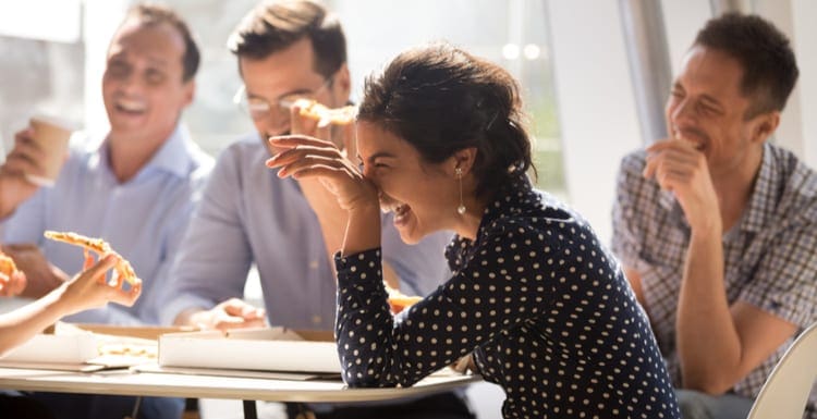 Indian woman laughing at funny real estate jokes eating pizza with diverse coworkers in office, friendly work team enjoying positive emotions and lunch together, happy colleagues staff group having fun at break