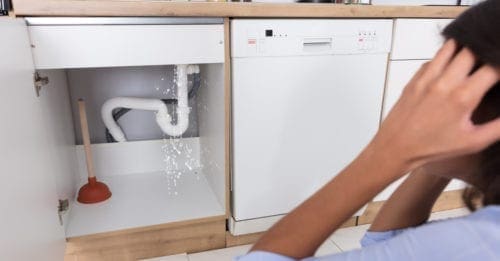 Someone standing holding their head and looking at their sink leaking through an open cabinet