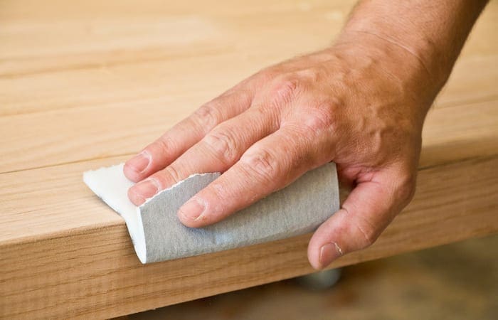 A middle aged hand holding a white piece of sandpaper and sanding an unfinished oak table
