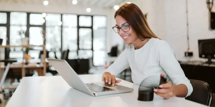 Smiling young woman using laptop and playing with slinky in bright office as the featured image for a piece on the pros and cons of being a real estate agent