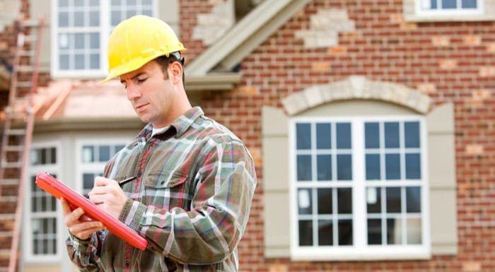 Man in a yellow hard hat stands outside of a beautiful brick home with a ladder in the back as the featured image for a piece on real estate jobs