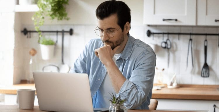 For a review on McKissock Learning, Pensive young Caucasian man sit at table at home kitchen work online on laptop pondering thinking. Thoughtful millennial male look at computer screen browsing wireless internet on modern gadget.
