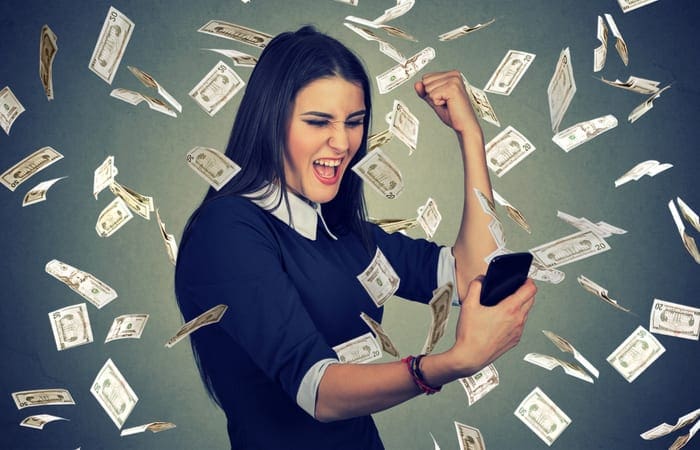 Successful happy woman using holding smart phone excited under money rain dollars falling down