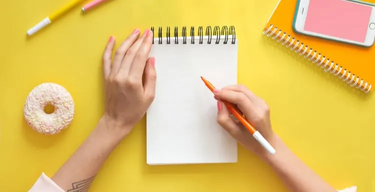 A featured image describing creating the perfect real estate bio. Colorful yellow background with stationary, donut, smart phone mock-up woman's hands writing in empty notebook. Flat lay top view. Learning to draw, making wish list or plans. Art education concept