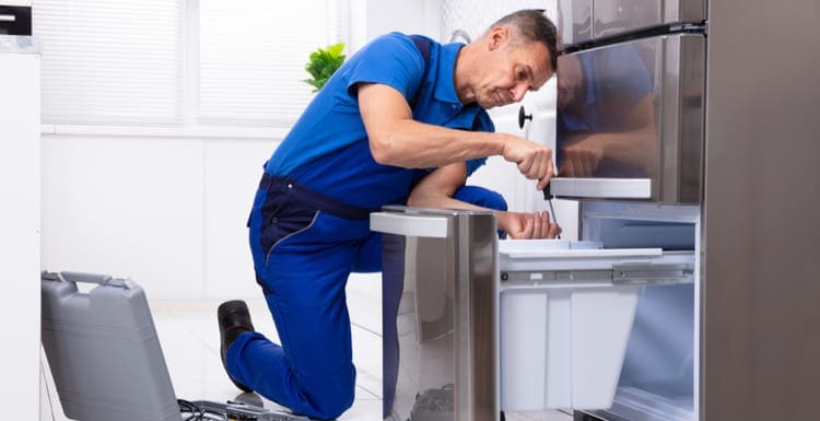 As a featured image for a piece on Refrigerator Repair Near Me, Mature Male Serviceman Repairing Refrigerator With Toolbox In Kitchen
