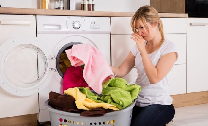 Young Woman Looking At Smelly Clothes Out Of Washing Machine In Kitchen