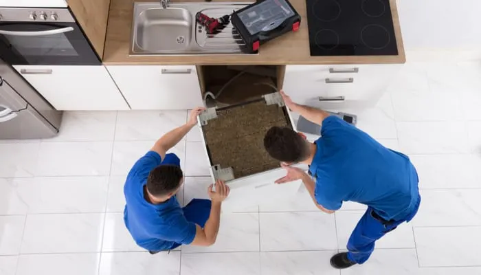 Two Young Male Movers In Uniform Placing Dishwasher In Kitchen