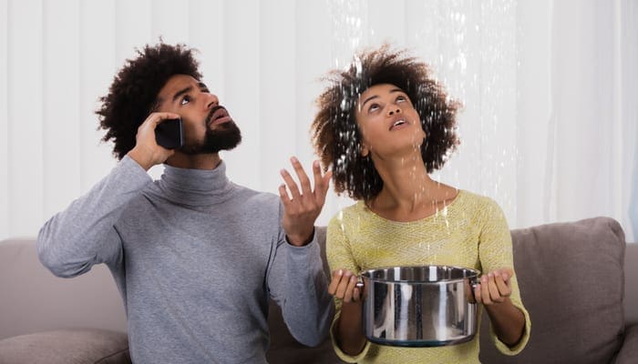 Young Man Calling Plumber While Woman Using Utensil For Collecting Water Leaking From Ceiling