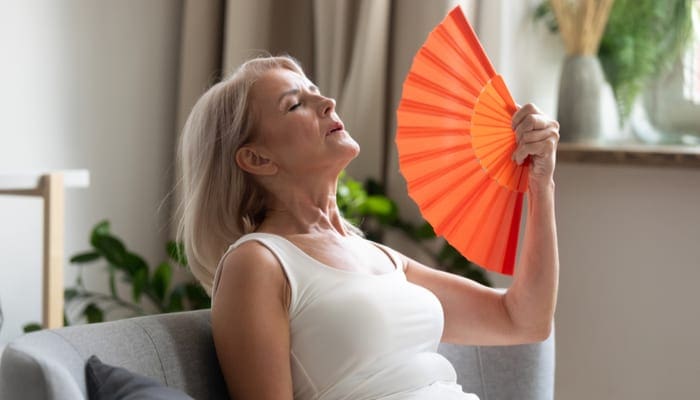 Stressed annoyed old senior woman using waving fan suffer from overheating, summer heat health hormone problem, no air conditioner at home sit on sofa feel exhaustion dehydration heatstroke concept
