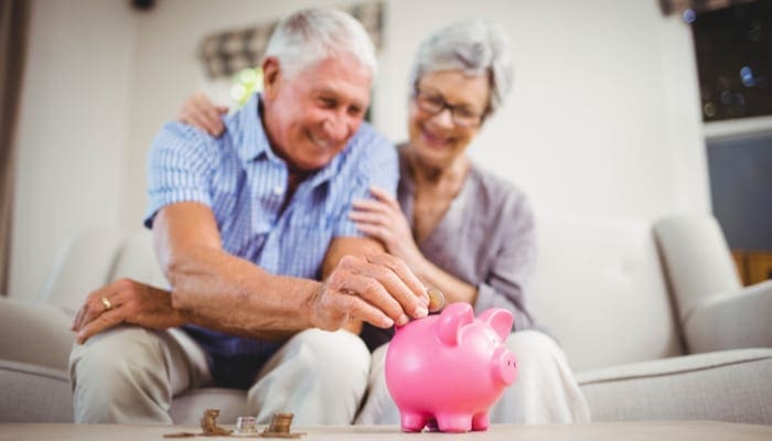 To symbolize how much a granny pod costs, senior man sitting with woman on sofa and putting coins in piggy bank