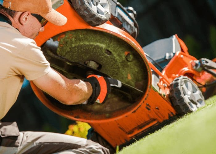 Below image of a man showing us how to remove a lawn mower blade with gloves and an orange hat on from below
