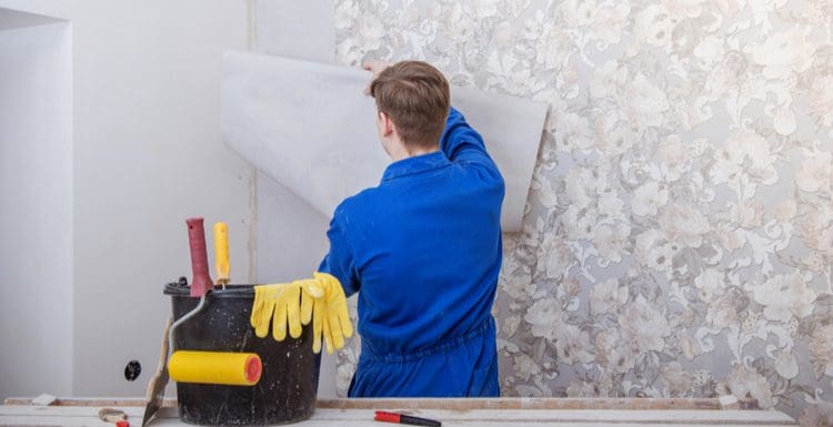 Featured image for a piece on Can You Paint Over Wallpaper featuring a man holding a piece of paper up to a swirly floral wall with a toolbox next to him