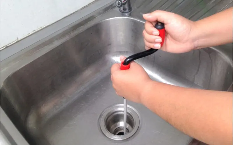 Man using a kitchen drain snake on a stainless steel sink bowl that is clogged