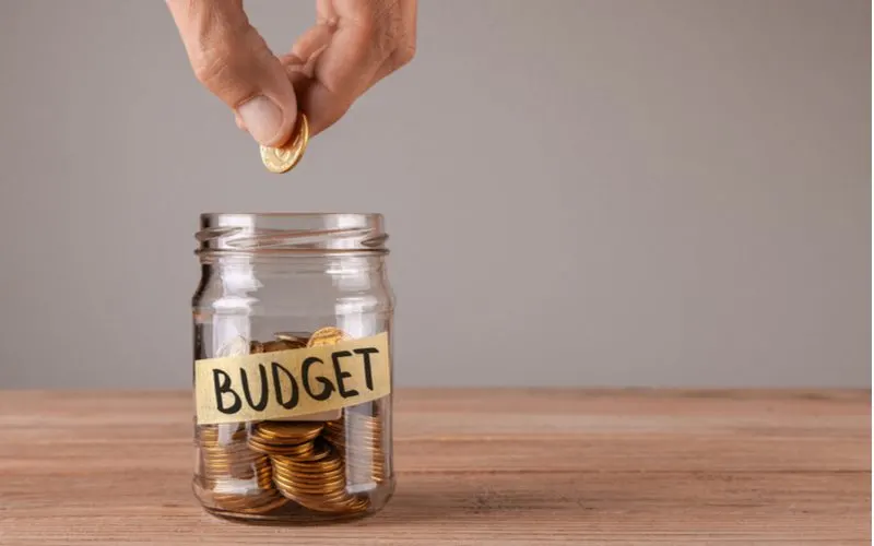 Man putting a coin into a glass jar titled Budget