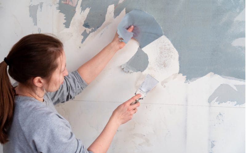 For a piece on can you paint over wallpaper, a woman with a paint scraper pulls down sections of the paper
