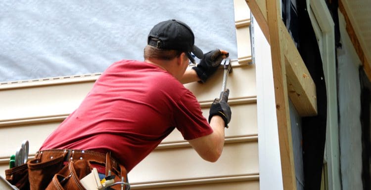 For a piece on the cost to side a house, a guy in a red shirt hammers a nail into a piece of vinyl siding