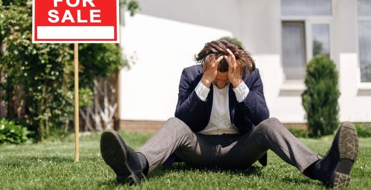 Image of a man sitting on a lawn in front of a for sale sign and holding his head for a piece on why do real estate agents fail
