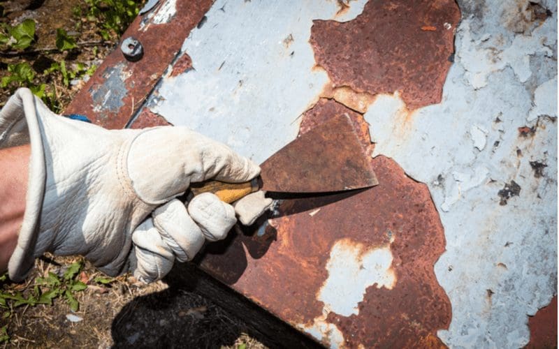 Man in white leather gloves removing paint from a metal sheet with a rusty paint scraper
