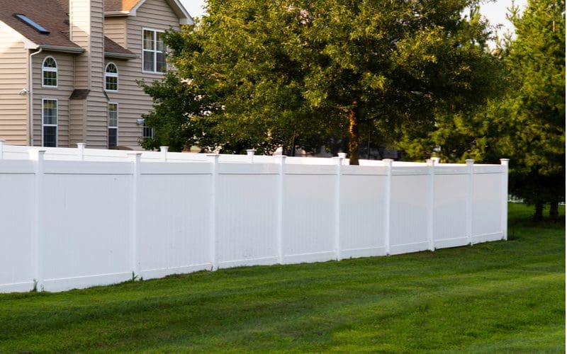 To illustrate factors affecting the vinyl fence price, a white vinyl fence in a suburban backyard next to green grass sits on a summer day