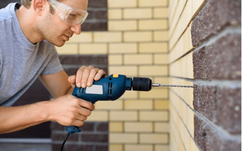 A man in goggles drills a brick wall with a drill
