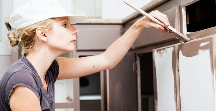Close-up of a woman painting laminate cabinets with brown paint and squatting down on her knees wearing a cap