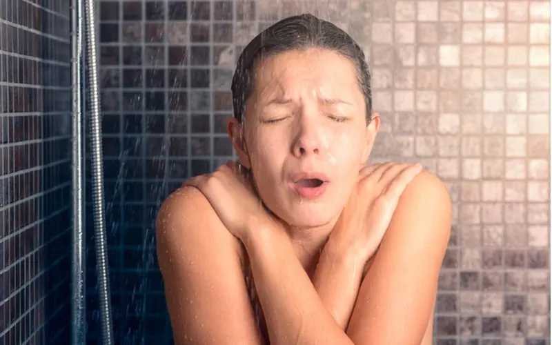 Bare chested woman covering her shoulders with her hands in an x shape and shivering after her water heater had problems
