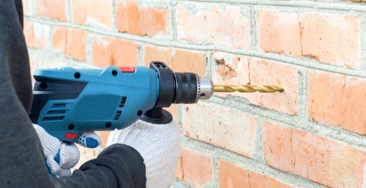 Image of a man drilling into brick with a green Makita drill while wearing gloves and a dark grey sweatshirt