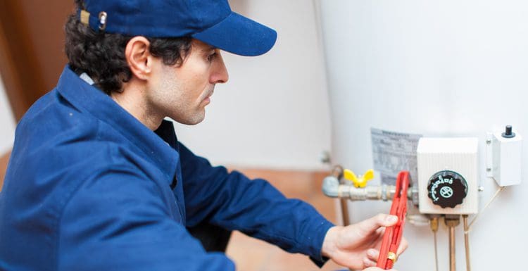 Featured image for a piece on hot water heater problems showing a man wrenching the burner assembly on a hot water heater and looking serious in a blue outfit with a blue hat
