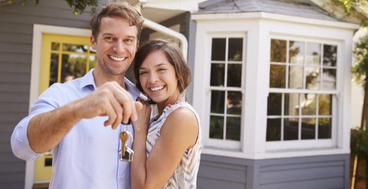 Cute couple standing outside of a grey house with a yellow door holding up the key for a piece on the best time to buy a house