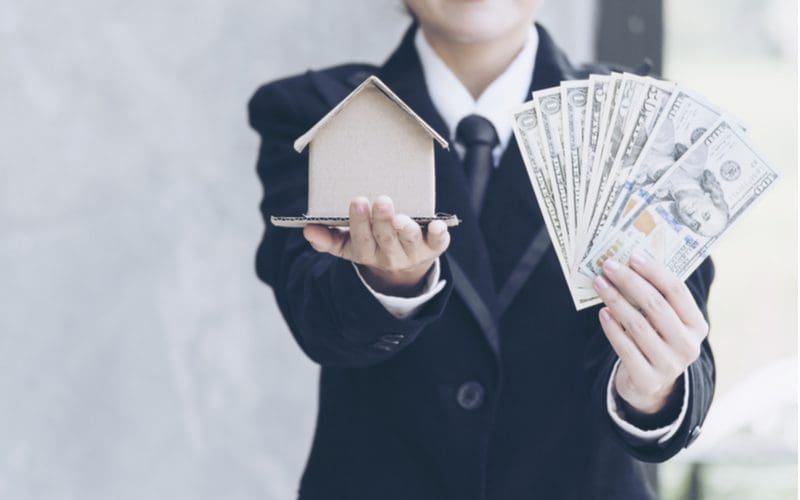 A man holding a wad of cash in one hand and a model house in another stands in a suit and you cannot see his face