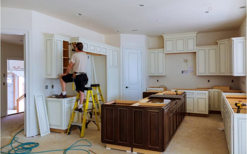 Man standing on a yellow step ladder remodeling a kitchen and nailing up white doors to cabinets