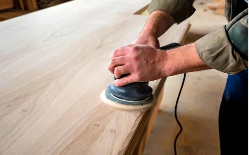 To show the reader how to paint pressure treated wood, a guy holding a random orbital sander and sanding the top of a piece of furniture
