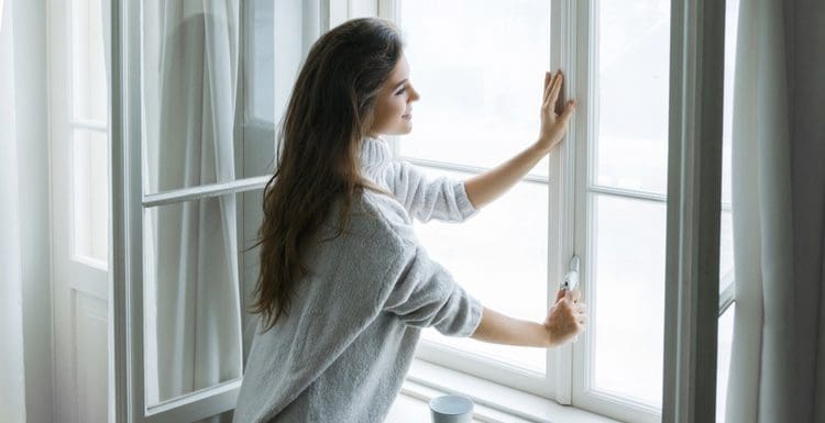 Woman cranking open a casement window while standing with a mug and wearing a cardigan sweater