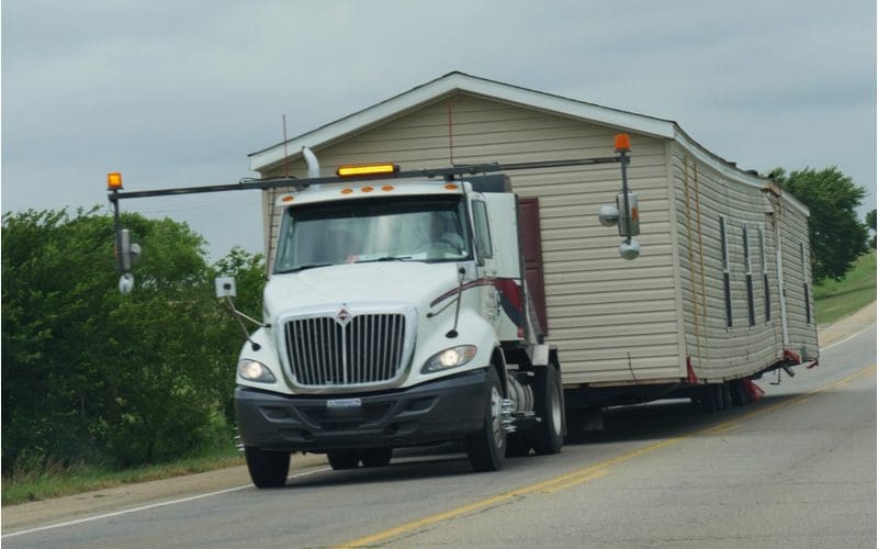 For a piece on how much does it cost to move a mobile home, a modular home sits on a trailer being pulled behind a white semi on a street with an oversized load light stack