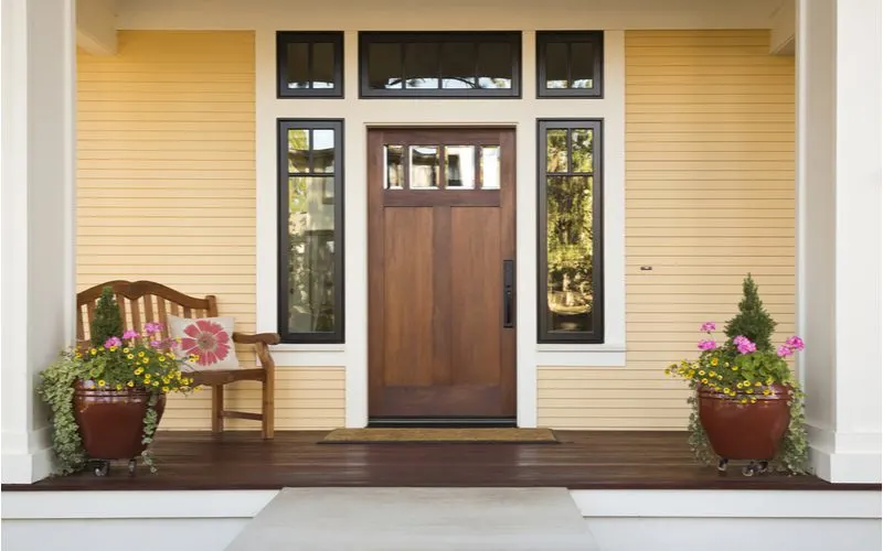 Large front door framed by two vertical windows and below three horizontal windows with yellow siding behind a rocking chair