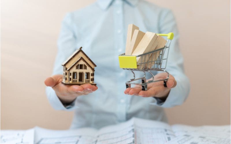Woman holding a small model house and a small shopping cart with small wooden blocks inside for a piece on is it cheaper to buy or build a house