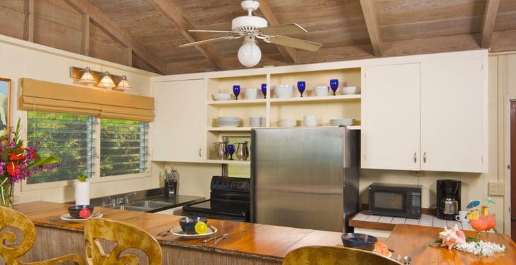 Featured image for a piece titled should you put a ceiling fan in kitchen, featuring a Hawaiian-style kitchen with wooden chairs and a fan on the ceiling