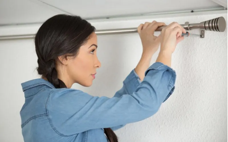 Woman handing a standard sized curtain rod on a stucco wall while wearing a blue shirt