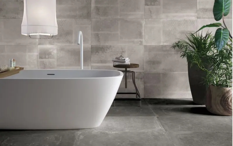 Wide grey floor tiles and multi-size wall grey ceramic wall tiles with an above-ground white ceramic tub with modern and simple minimalistic fixtures