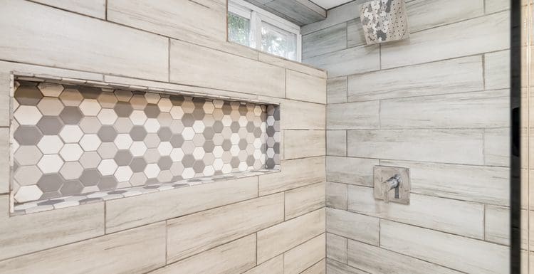 Featured image for a piece on tile shower ideas featuring a grey wood-look horizontal tile shower with a small hex tile patterned soap shelf and chrome fixtures